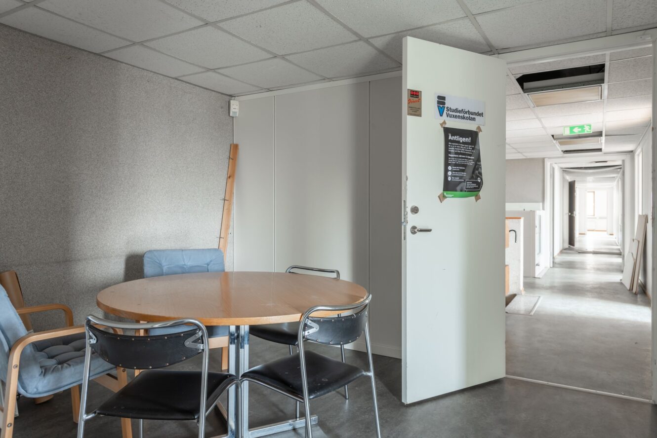 Ett litet fikarum med ett runt träbord, fyra stolar och en öppen dörr som leder ut till en korridor med synliga takpaneler och lysrörsbelysning.