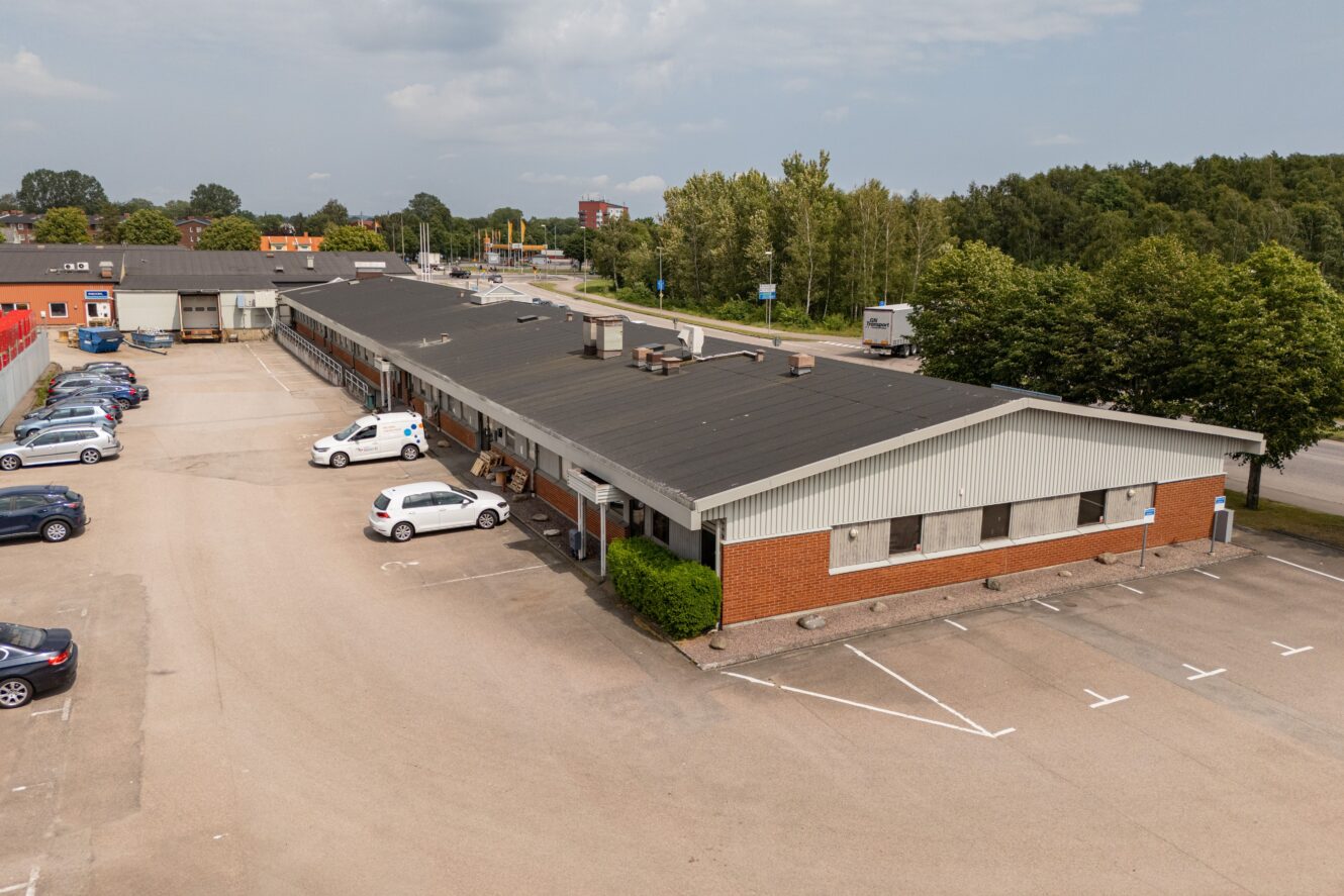 En envånings industribyggnad med en stor parkeringsplats, flera parkerade bilar och träd i bakgrunden under en delvis molnig himmel.