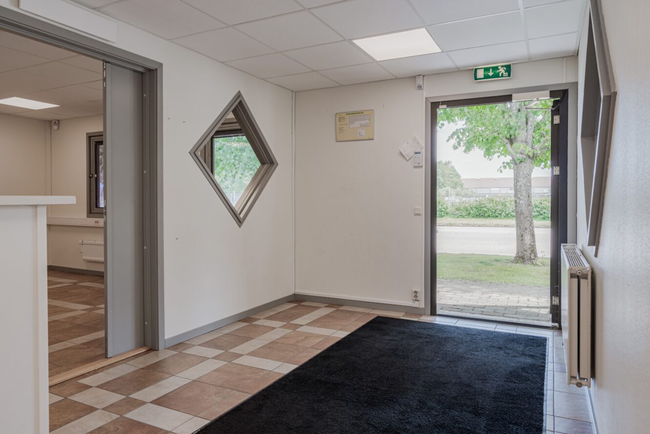 Tom kontorsentré med klinkergolv, svart matta, vita väggar, öppen dörr utåt och diamantformat fönster bredvid innerdörren.