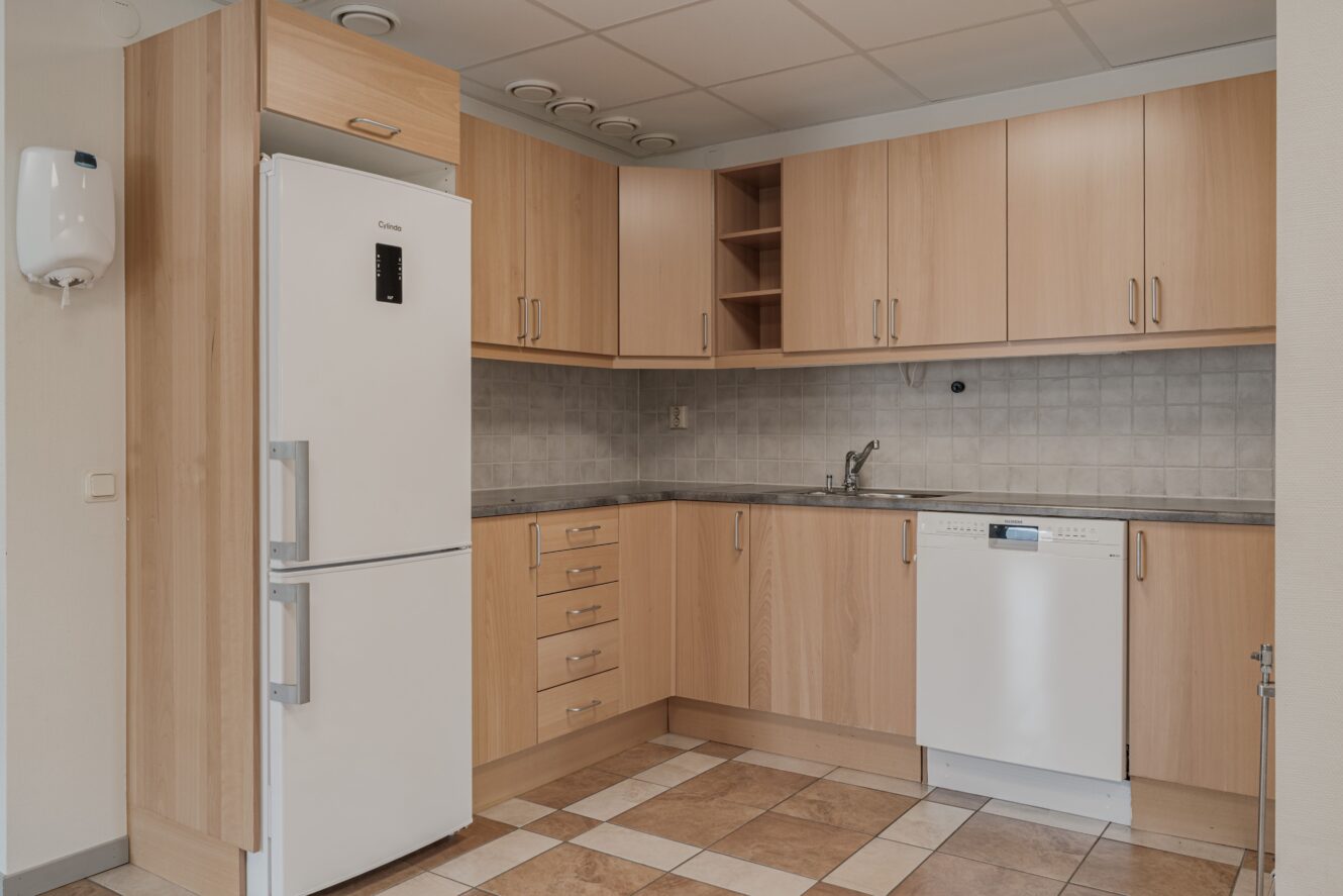 Modernt kök med skåp i ljust trä, vitt kylskåp, inbyggd diskmaskin, kaklat stänkskydd och klinkergolv. Diskbänk och bänkskiva finns i hörnet.