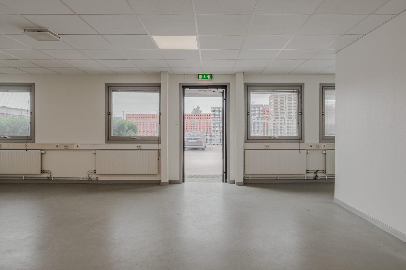 Tomt rum med grått golv, vita väggar, tre fönster och en öppen dörr som leder ut till en parkeringsplats med byggnader som syns i bakgrunden.