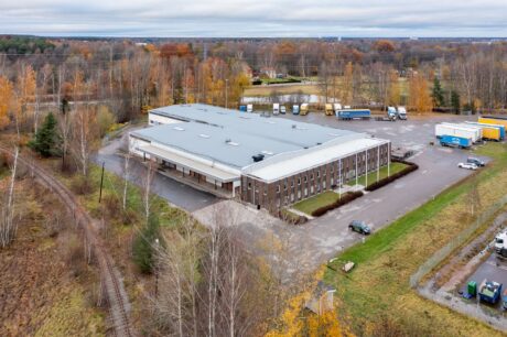 Flygfoto över ett stort industrilager med lastkajer, omgivet av träd och lastbilar, beläget nära ett järnvägsspår i ett höstlandskap.