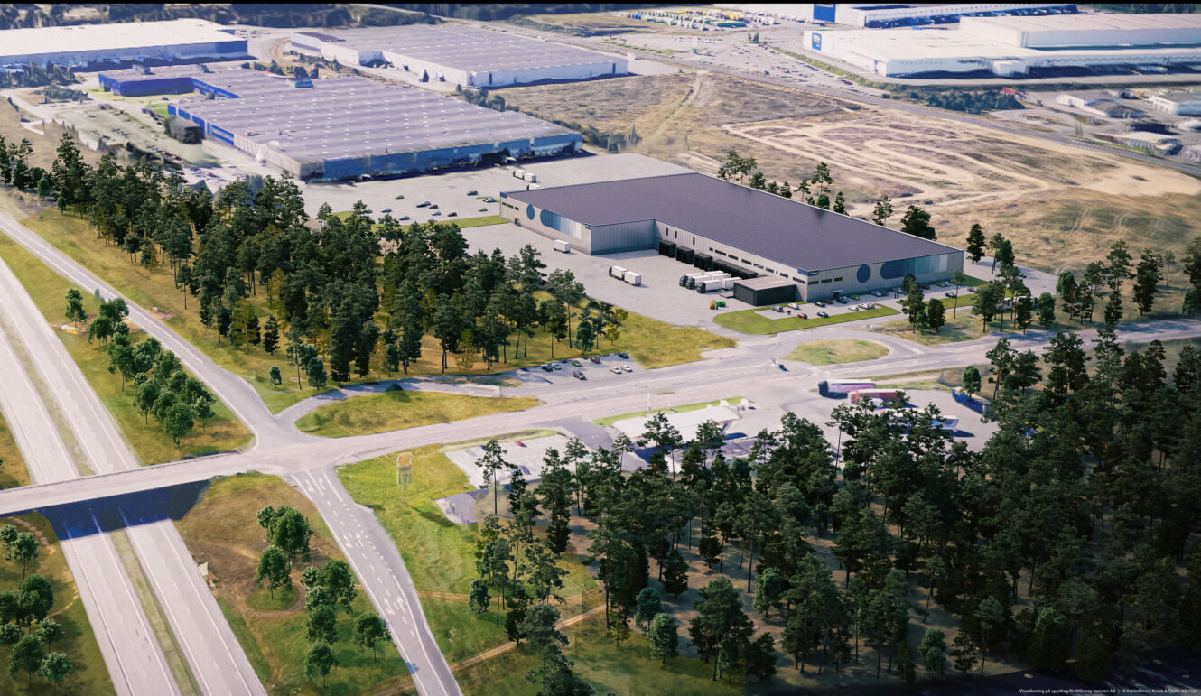 Flygfoto över ett industriområde med stora lagerlokaler, parkeringsplatser, lastbilar och omgivande vägar, kantat av träd och skogsmark.