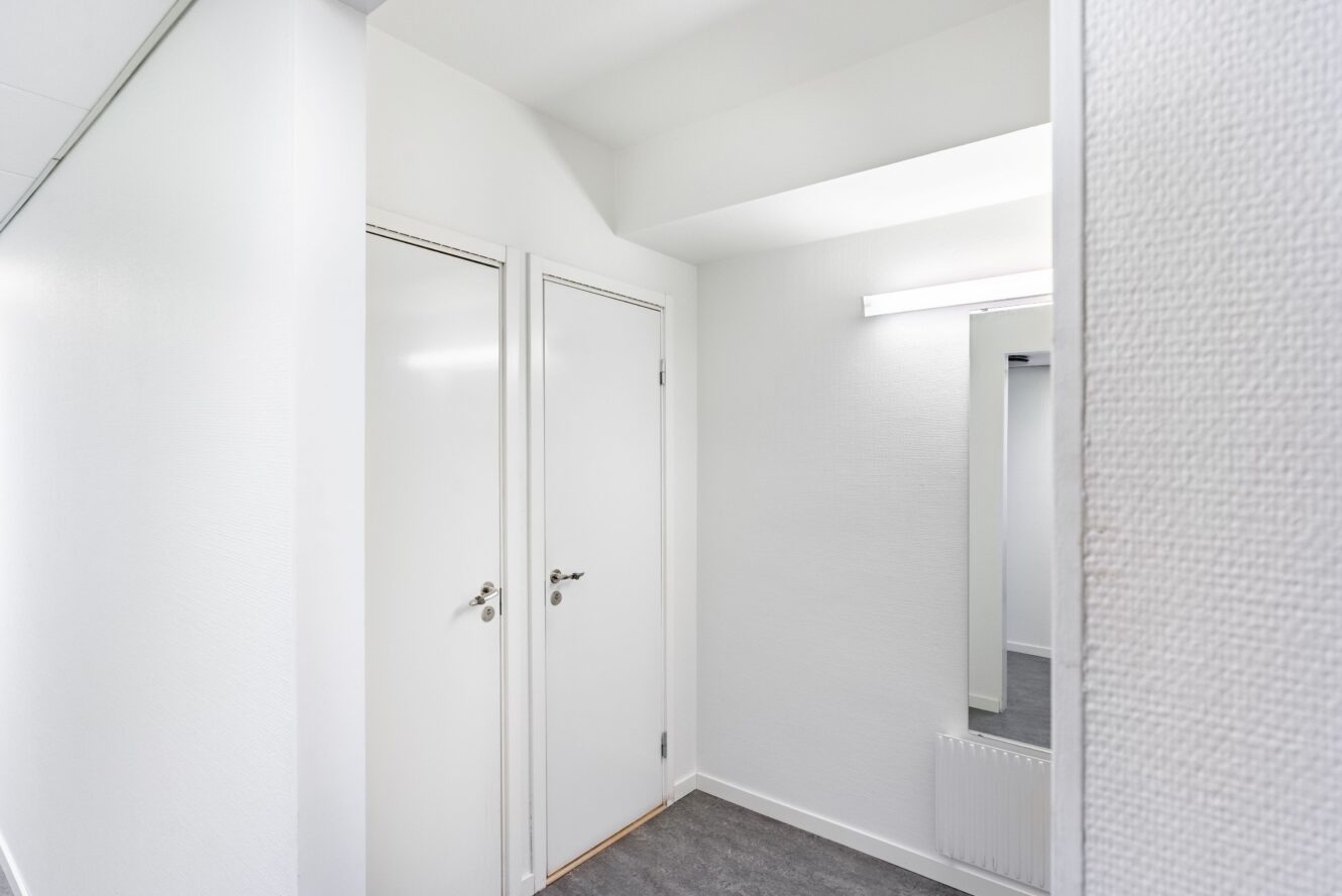 En korridor med två stängda vita dörrar till vänster, en väggmonterad spegel och ett lysrör ovanför spegeln.