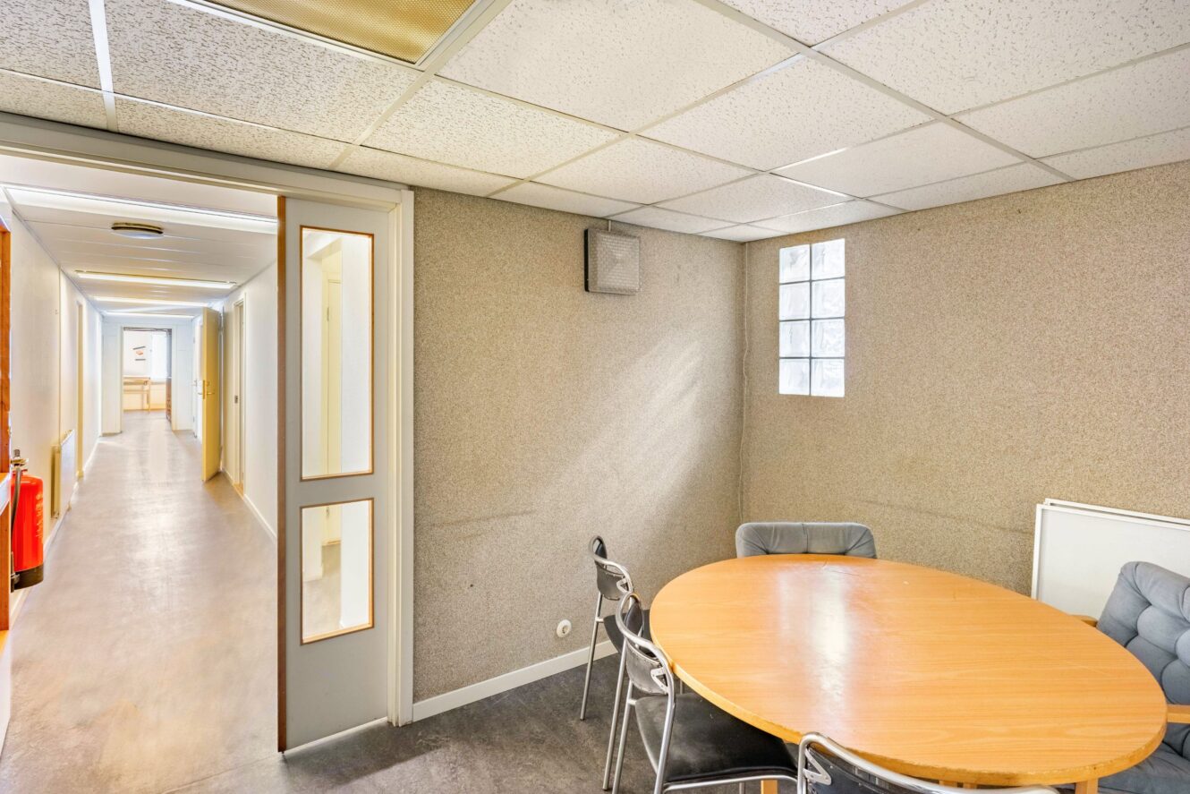 Litet mötesrum med ett runt träbord och fyra stolar, intill en korridor med vita väggar och takplattor. Naturligt ljus kommer in genom ett fönster med glasblock.