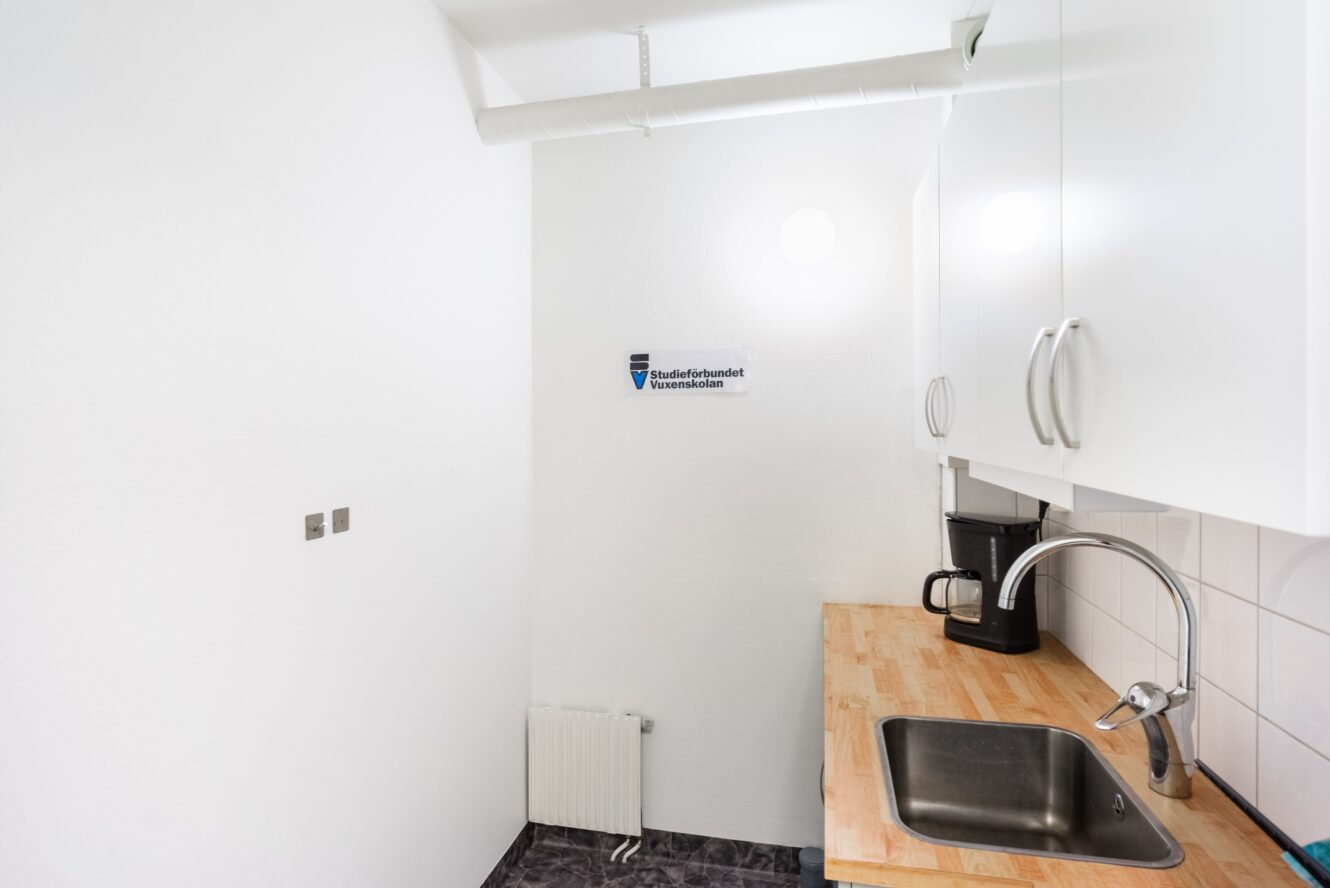 Litet pentry med vita skåp, bänkskiva i trä, diskho, kaffebryggare, vitt kakel och ett element under en skylt på väggen.