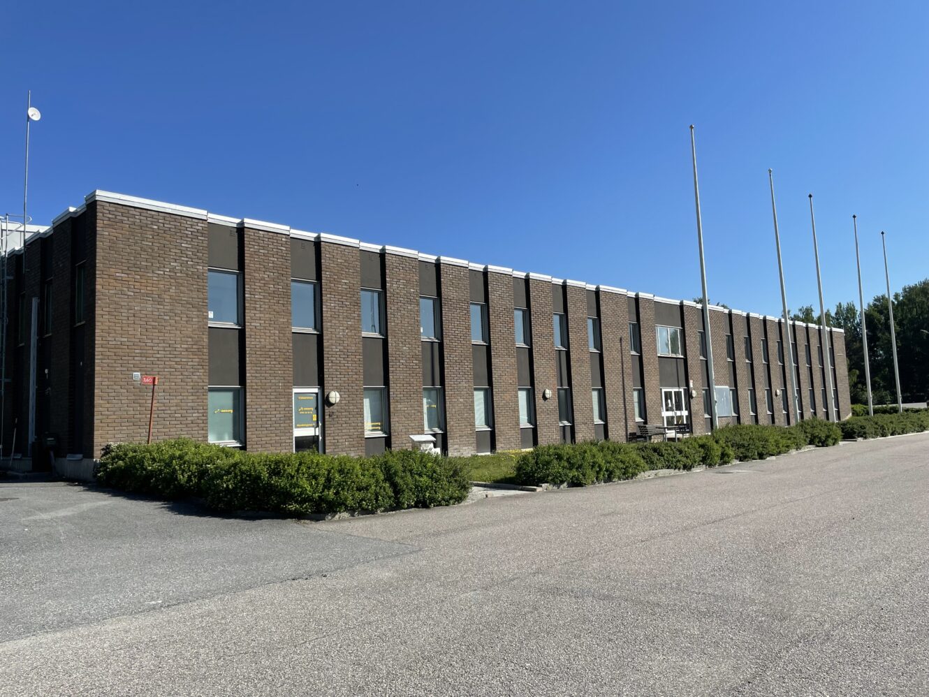 En tvåvånings kontorsbyggnad i tegel med rader av fönster, omgiven av buskar och tomma flaggstänger, under en klarblå himmel.