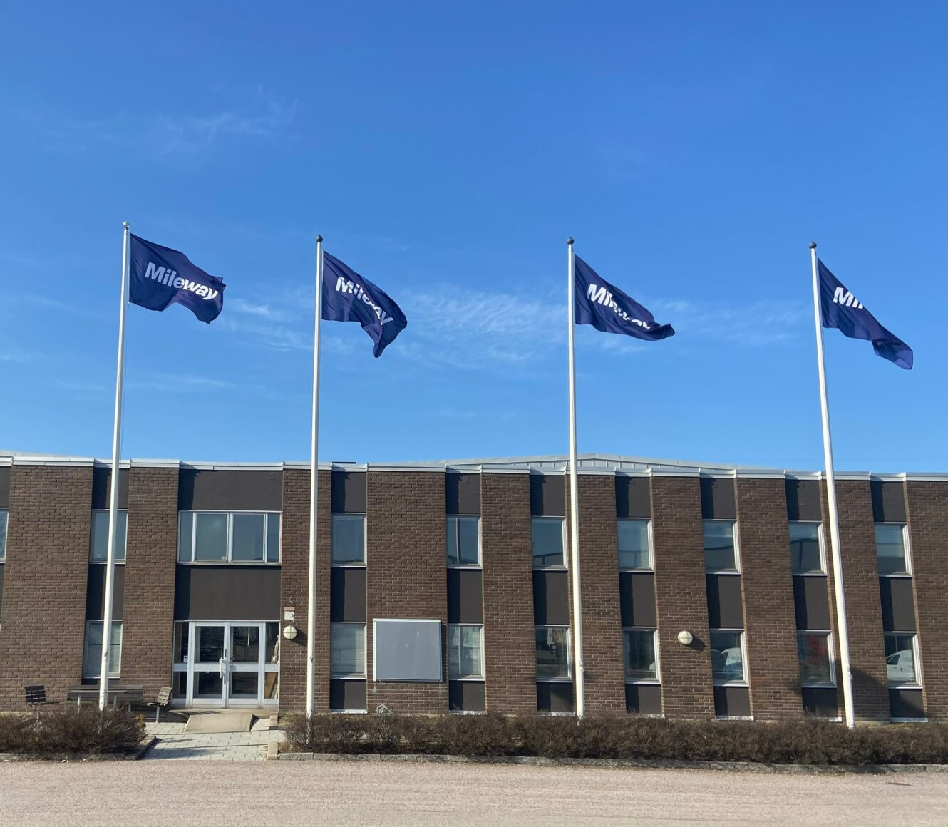 En kontorsbyggnad i tegel med fyra flaggstänger med blå flaggor där det står Milwau under en klarblå himmel.