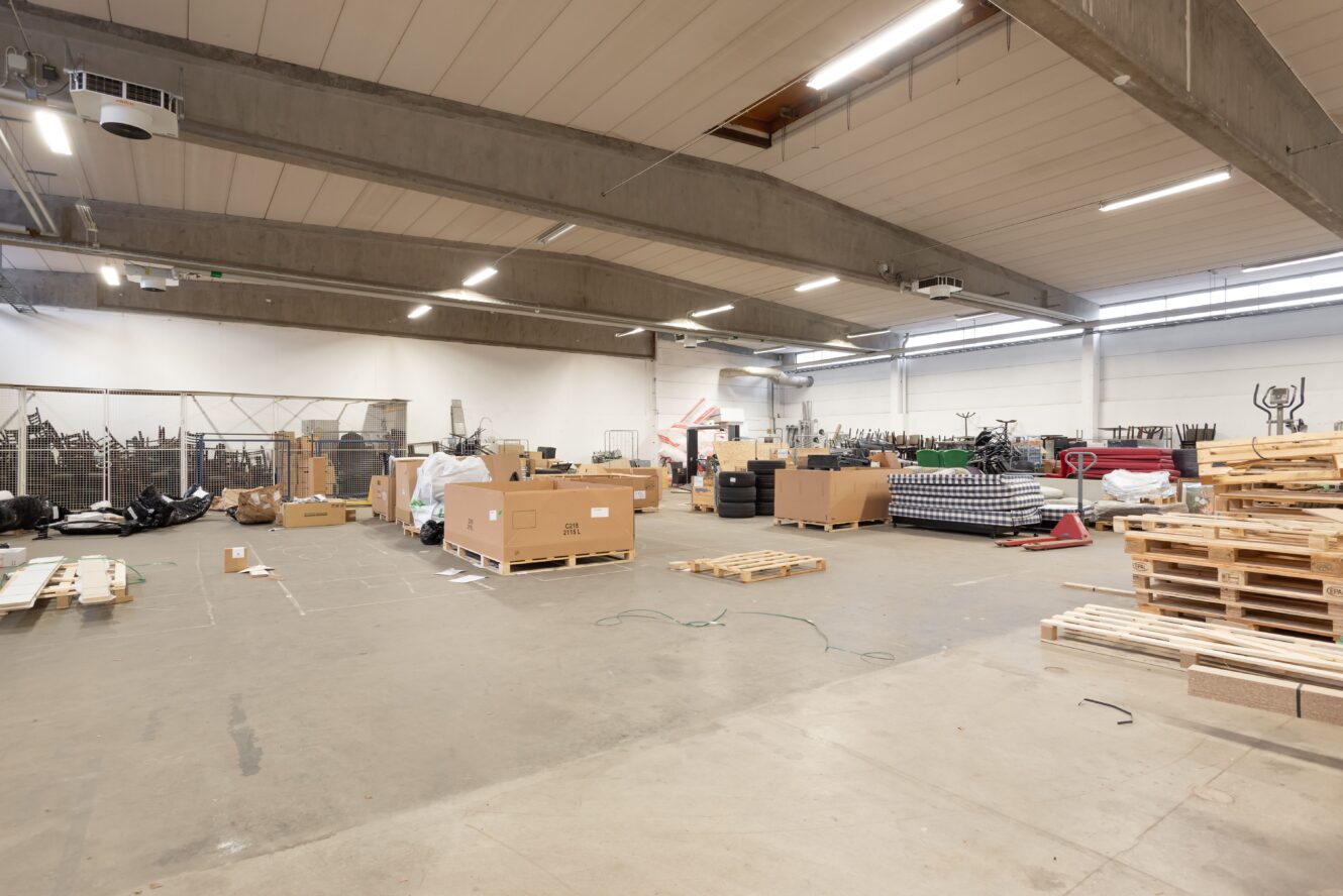 En rymlig lagerlokal med högt i tak, utspridda lastpallar, kartonger, trälådor och olika material organiserade längs väggarna.