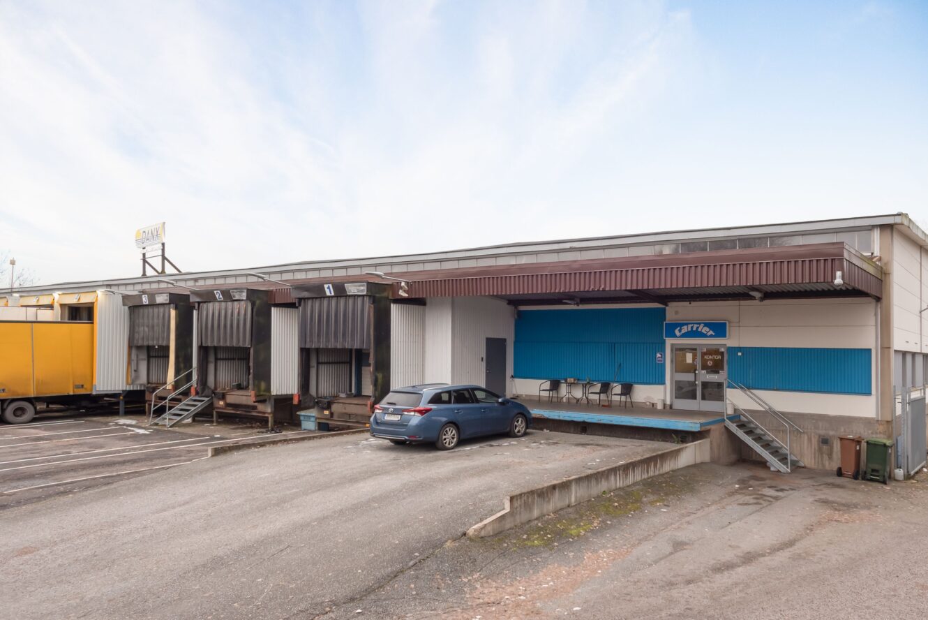 En liten industribyggnad med tre lastkajer, en gul lastbil, en parkerad bil och en ingång med texten Cafeteria på höger sida.