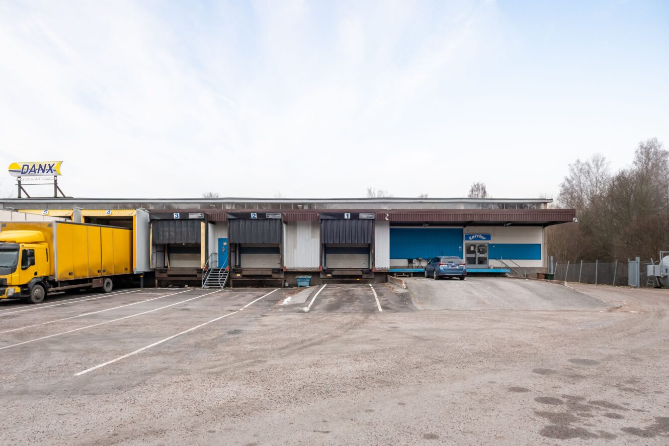 Ett distributionslager med fem lastbryggor, en gul lastbil parkerad till vänster, en blå bil till höger och en tom parkeringsplats framför.