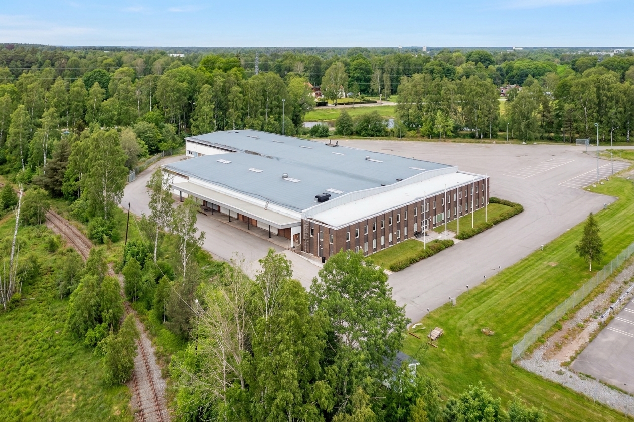 En stor, rektangulär lagerbyggnad med grått tak omges av grönska, en parkeringsplats och träd, sett uppifrån en klar dag.