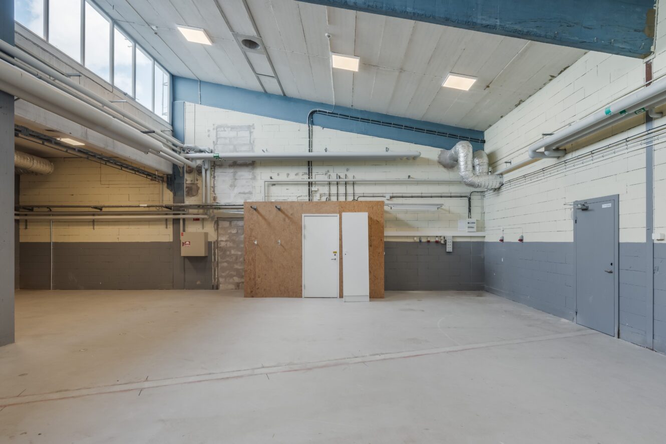 Rymligt, tomt industrirum med högt i tak, synliga rör, en liten träkonstruktion i mitten och gråmålade väggar.