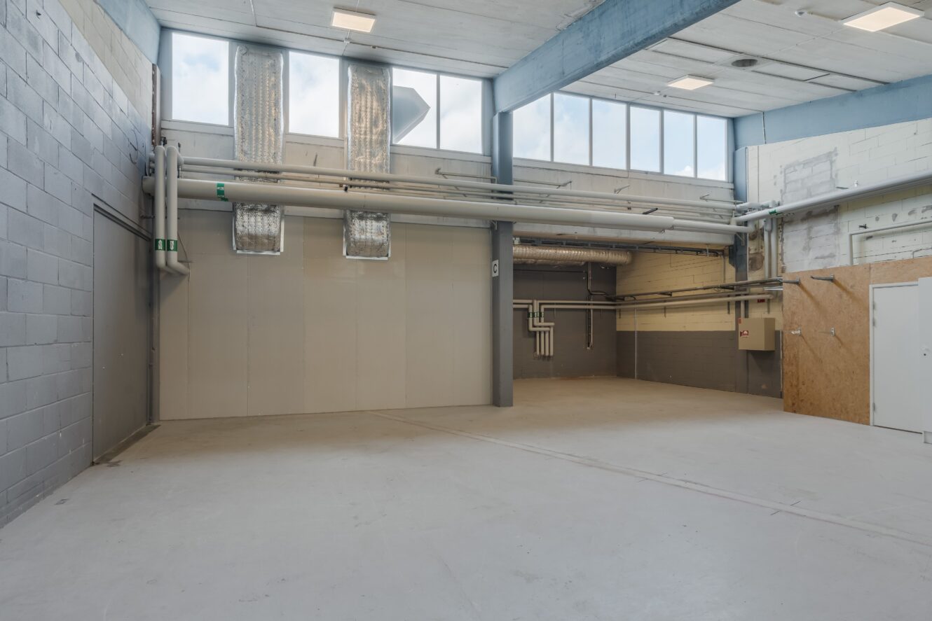 Tomt industriellt lagerutrymme med högt i tak, synliga rör, kanalsystem, stora fönster och betonggolv.