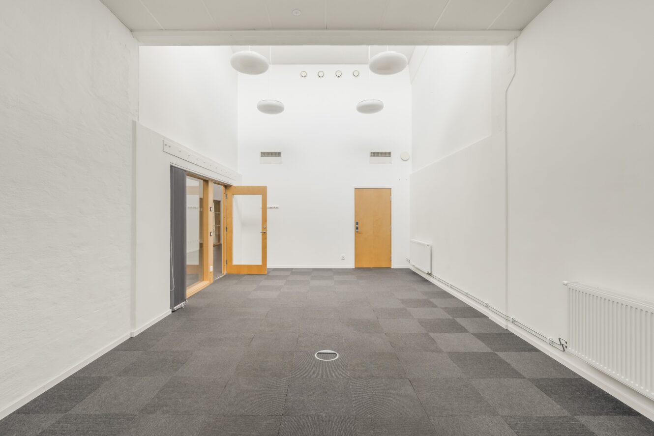 Ett rymligt, tomt rum med vita väggar, grå heltäckningsmatta, tre trädörrar och stora runda taklampor.