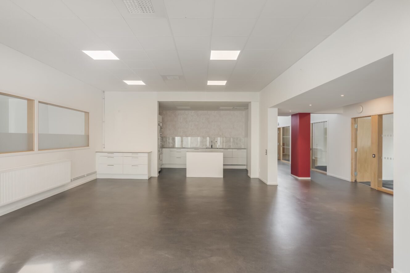 Ett rymligt, tomt rum med vita väggar, stora fönster, takbelysning och ett pentry längst bak. Golvet är mörkt och det finns öppna dörröppningar till angränsande rum.