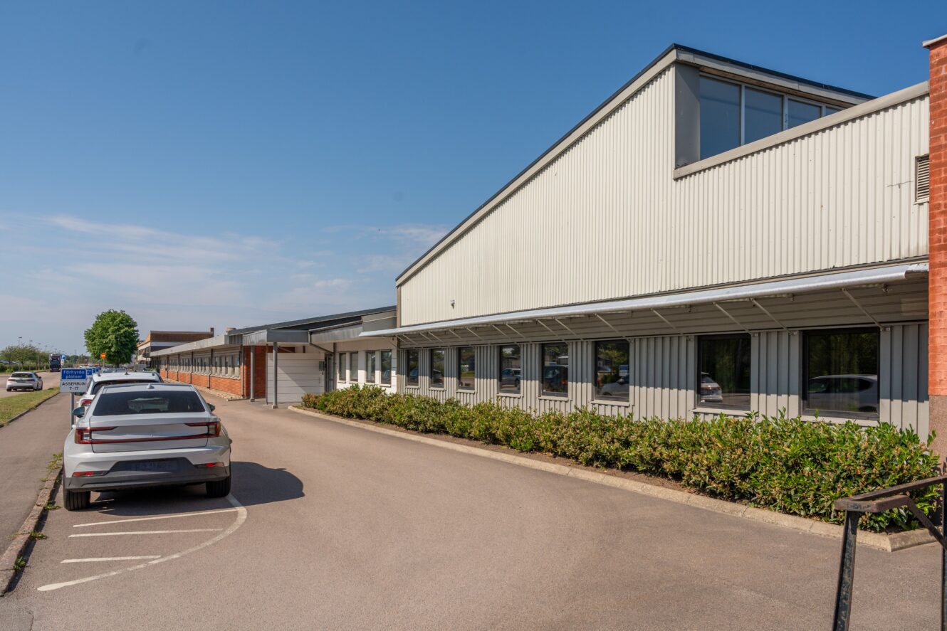 En stor industri- eller affärsbyggnad med vit metallfasad, flera fönster och parkerade bilar längs en asfalterad uppfart en klar dag.