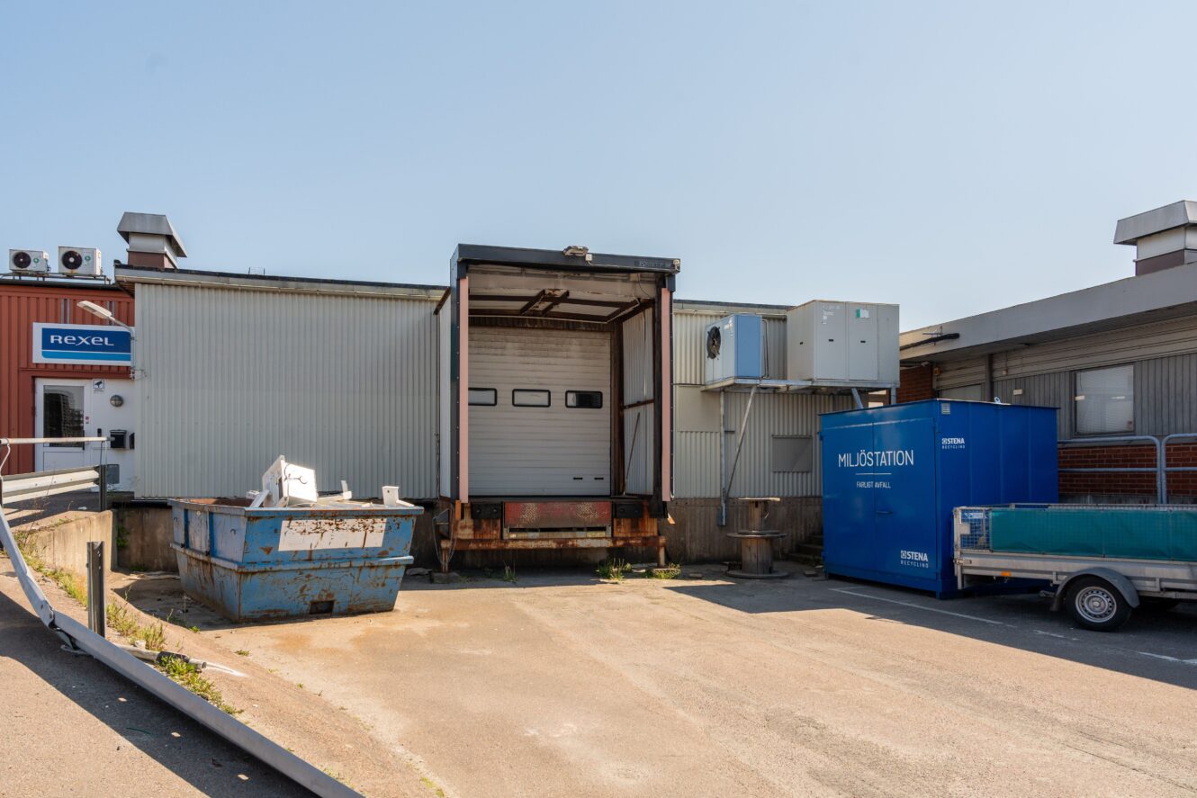 En lastkaj med stängd takskjutport, en blå container till vänster och en stor blå återvinningsstation till höger. Området ser tomt och industriellt ut.