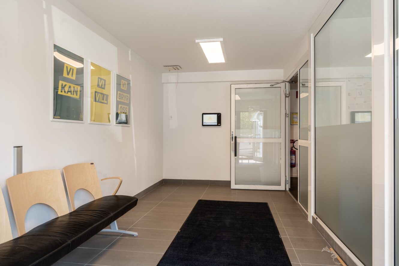 Ett litet väntrum med två trästolar, inramade affischer på väggen, klinkergolv, en svart matta och en glasdörr som leder till ett annat rum.