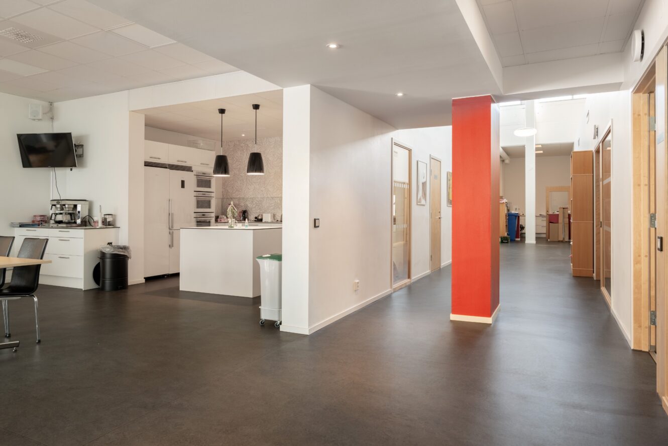 Modern kontorsinredning med pentry, vita väggar, en röd accentpelare, kontorsdörrar i glas och taklampor.