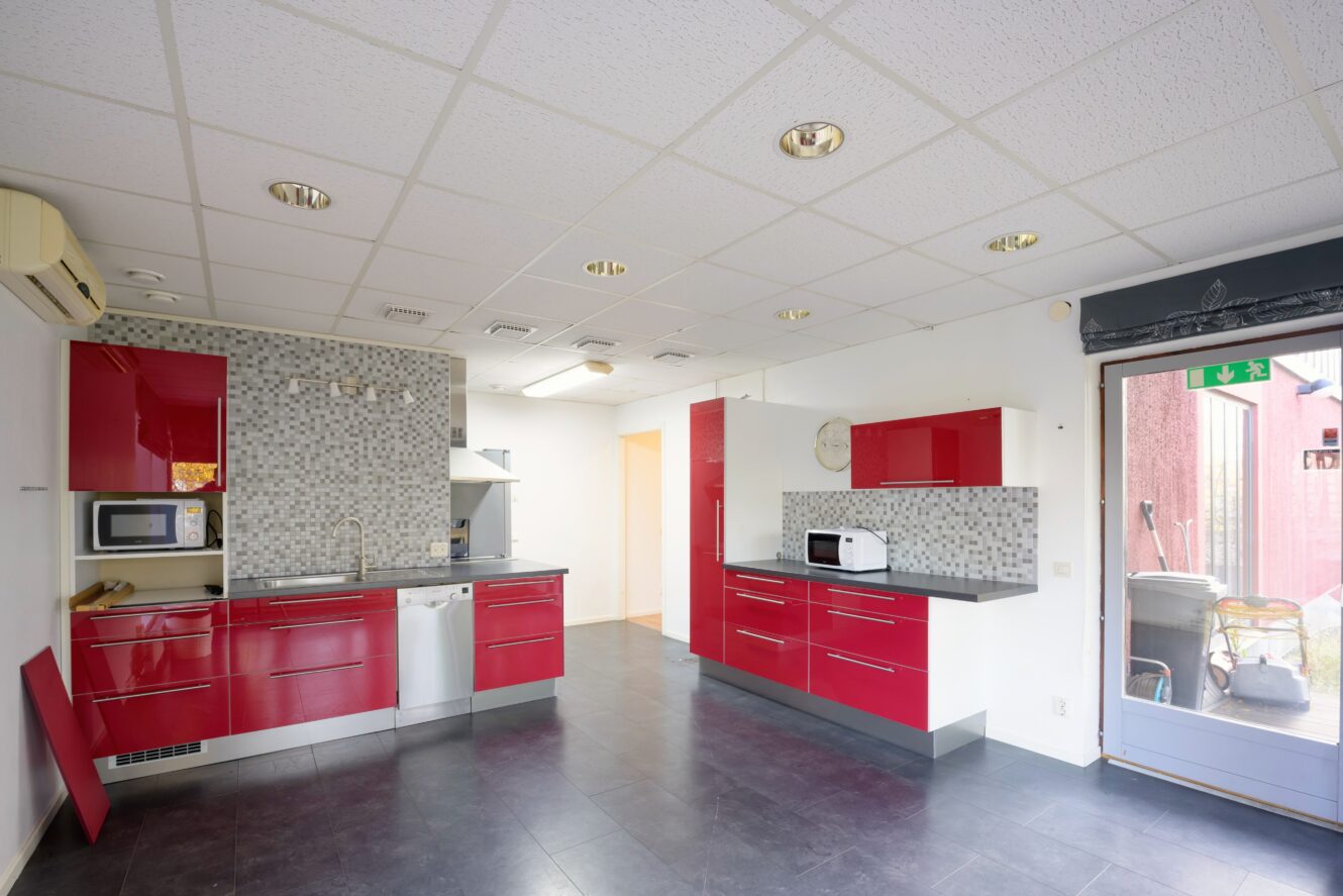 Modernt kök med röda skåp, grått kaklat stänkskydd, inbyggda vitvaror, mikrovågsugn på bänkskivan och en dörr som leder ut.