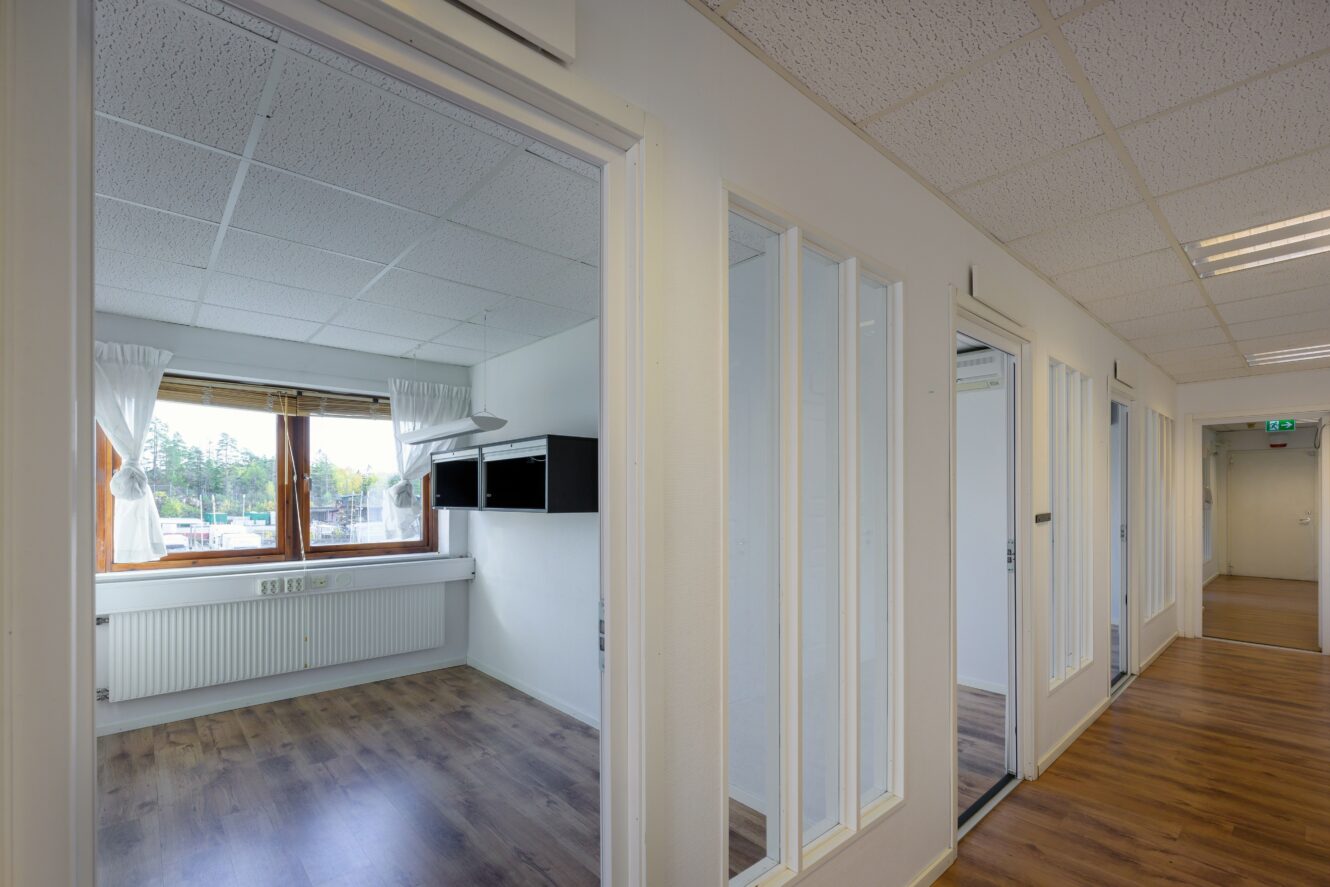 En korridor med dörrar som leder till tomma kontorsrum, med stora fönster, vita väggar, trägolv och taklampor.