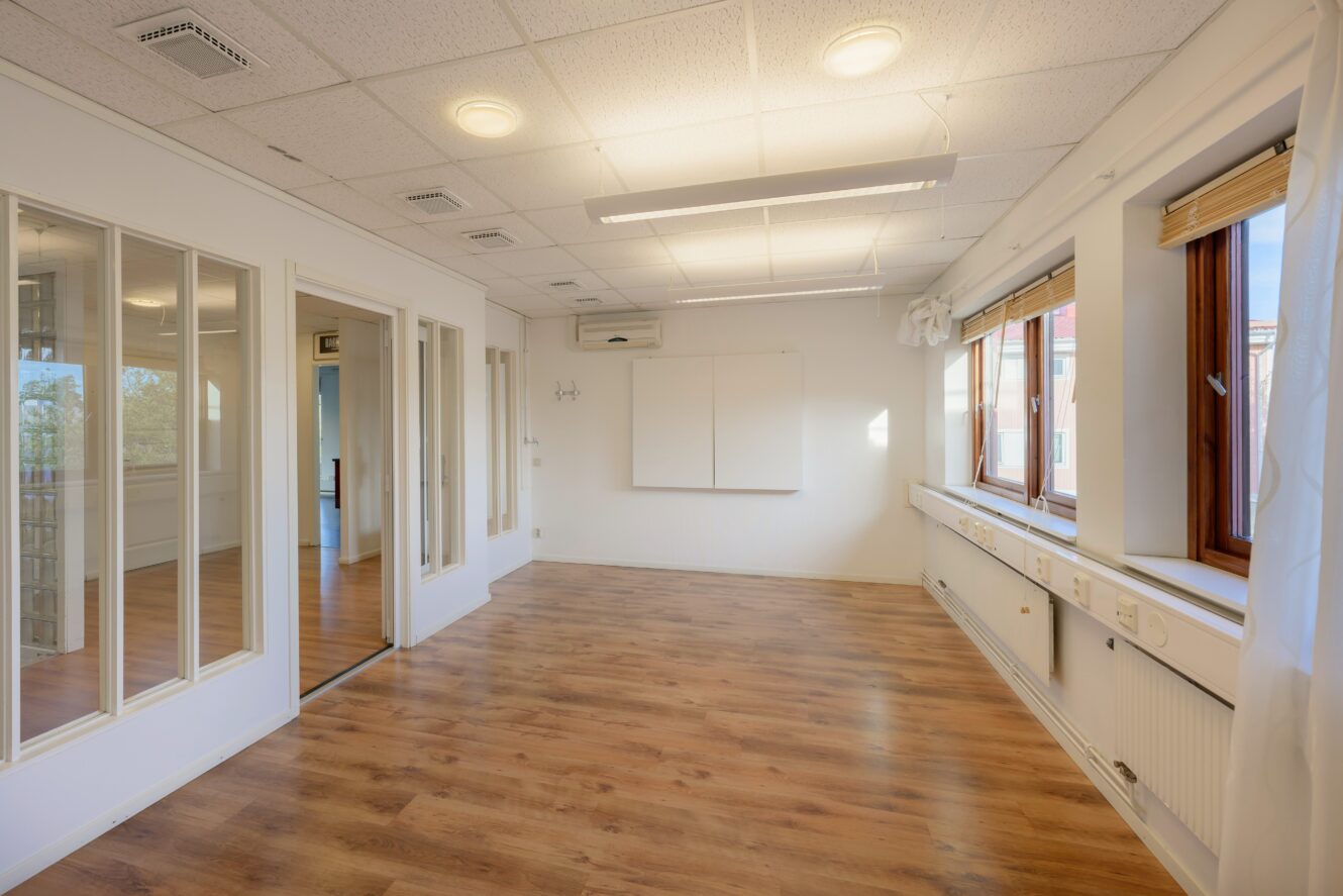 Ett tomt rum med trägolv, vita väggar, stora fönster på ena sidan, glaspartier och taklampor.