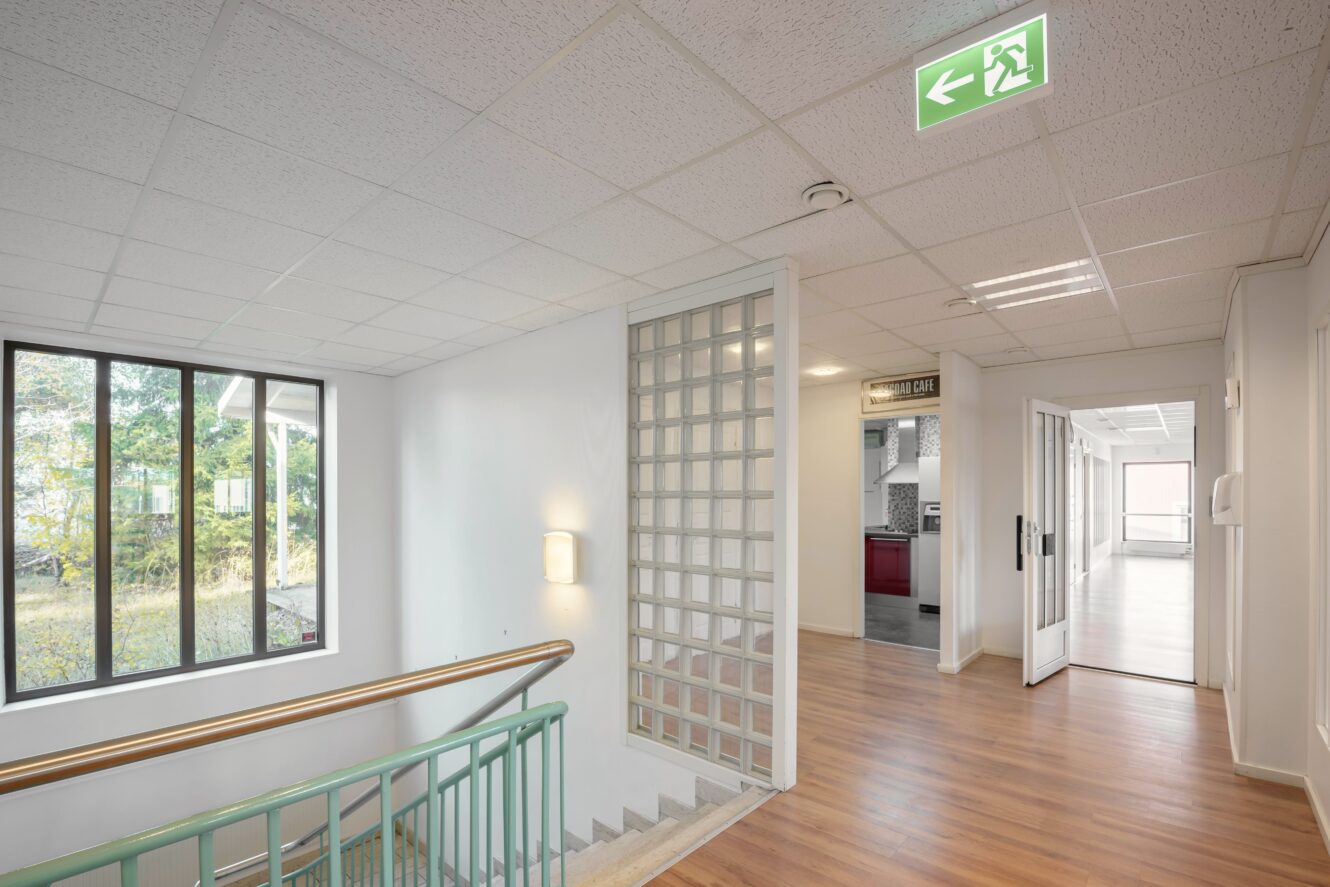 En modern hall med trägolv, grönt räcke vid trappan, glasvägg, stort fönster och en grön nödutgångsskylt i taket.