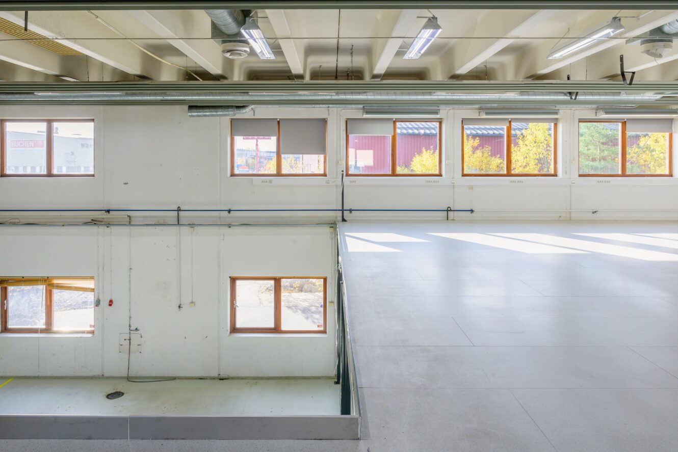 Inredning i en industribyggnad med stora fönster, vita väggar, synliga rör och en våning i två plan med gott om naturligt ljus.