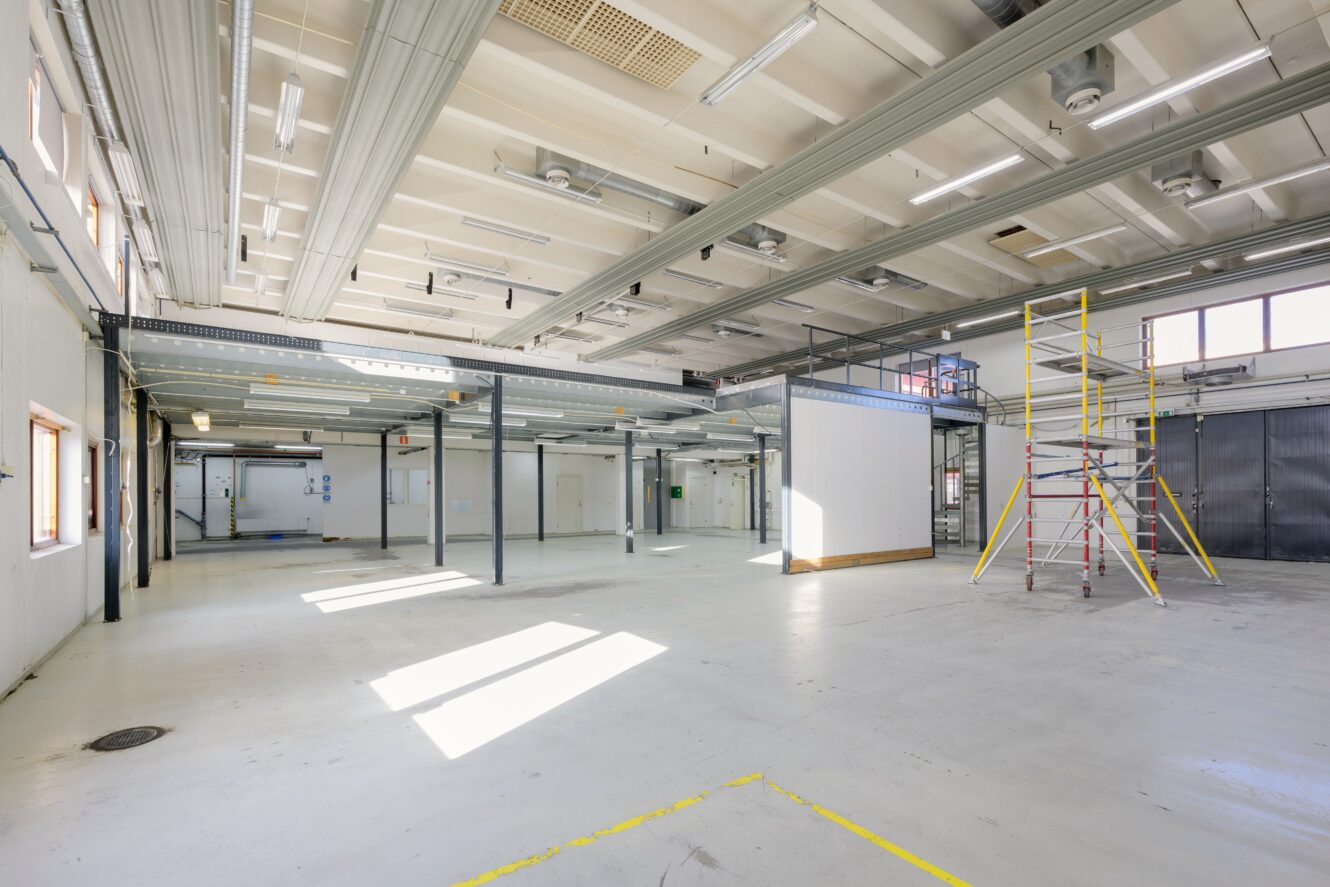 Rymlig, väl upplyst industriinteriör med vita väggar, synliga bjälkar, upphöjda plattformar och en byggnadsställning nära mitten. Solljuset flödar in genom fönstren till vänster.