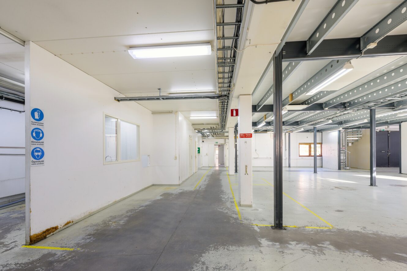 Ett tomt industrilager med vita väggar, stödbalkar av metall, takbelysning, säkerhetsskyltar och markerade gångvägar på golvet.