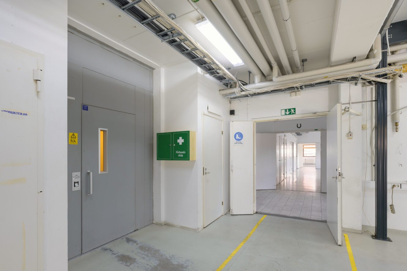 Industrihall med en grå hiss, första hjälpen-låda, synliga rör och en öppen dörr som leder till en starkt upplyst korridor.