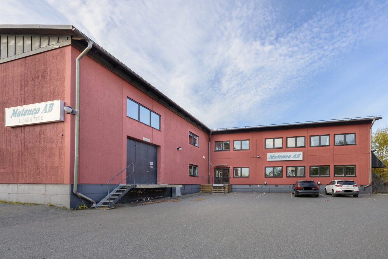 En röd industribyggnad i två våningar med Matenco AB Logistics-skyltar, en lastkaj och en parkeringsplats med två bilar.
