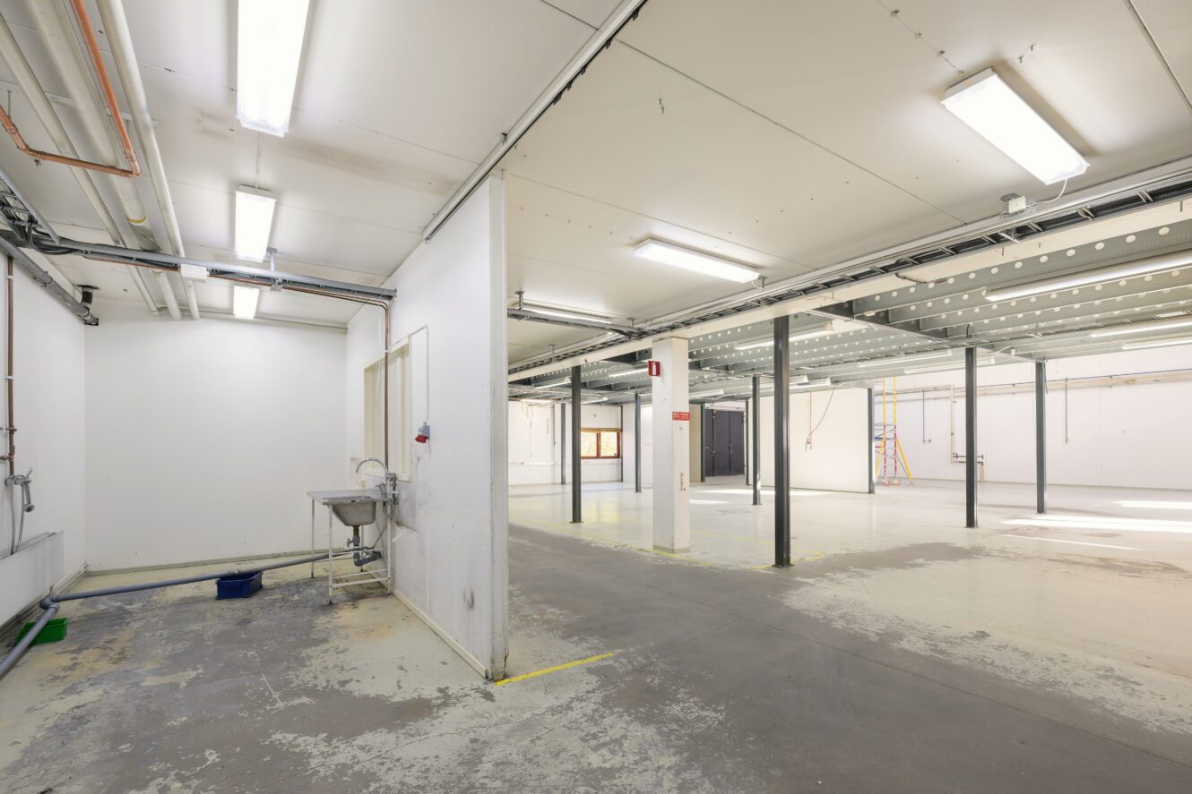 Stor tom industrilokal med vita väggar, synliga tak, metallpelare, lysrör i taket och en diskho till vänster. Golvet visar tecken på slitage.
