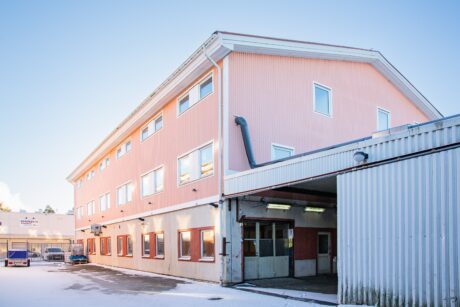 En trevånings rosa industribyggnad med flera fönster och en tillhörande korrugerad metallstruktur, omgiven av snötäckt mark en klar dag.