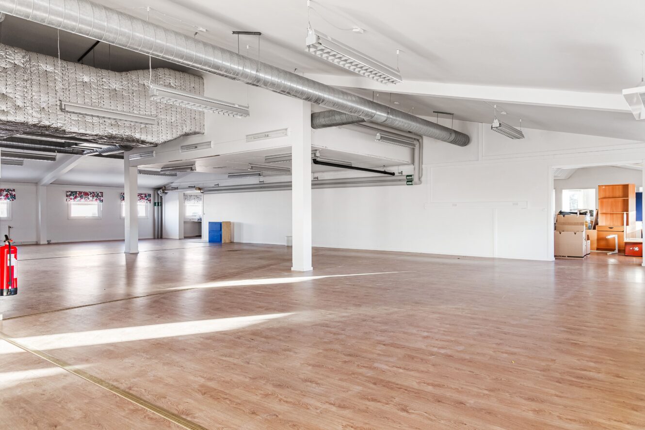 Rymligt, tomt rum med trägolv, synliga kanaler i taket och några lådor och en brandsläckare vid ena väggen. Naturligt ljus kommer in genom flera fönster.