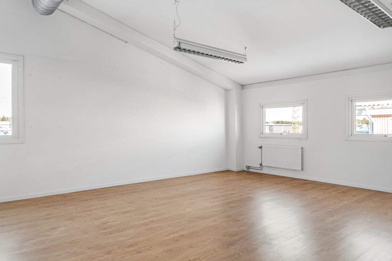 Ett stort, tomt rum med vita väggar, ljust trägolv, två fönster, ett element och lysrör i taket.
