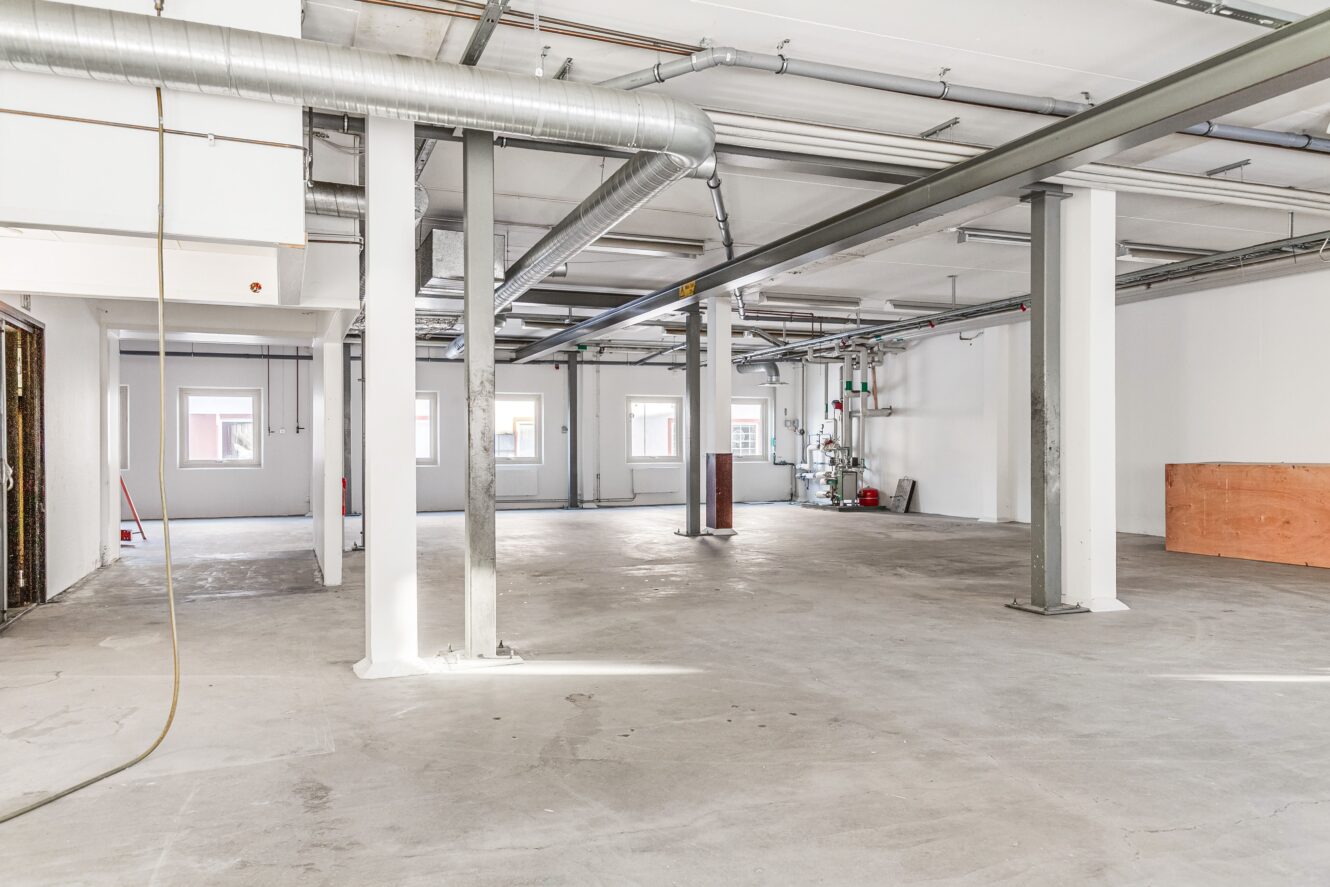 Interiör i en tom industrilokal med synliga rör, pelare och betonggolv; vita väggar och flera fönster ger naturligt ljus.