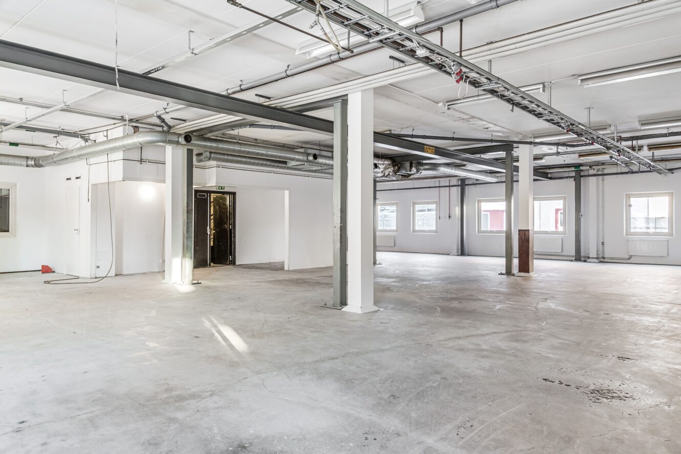 Stor, tom industrilokal med synliga rör, vita väggar och flera fönster som släpper in naturligt ljus; betonggolv och minimalt med inventarier syns.