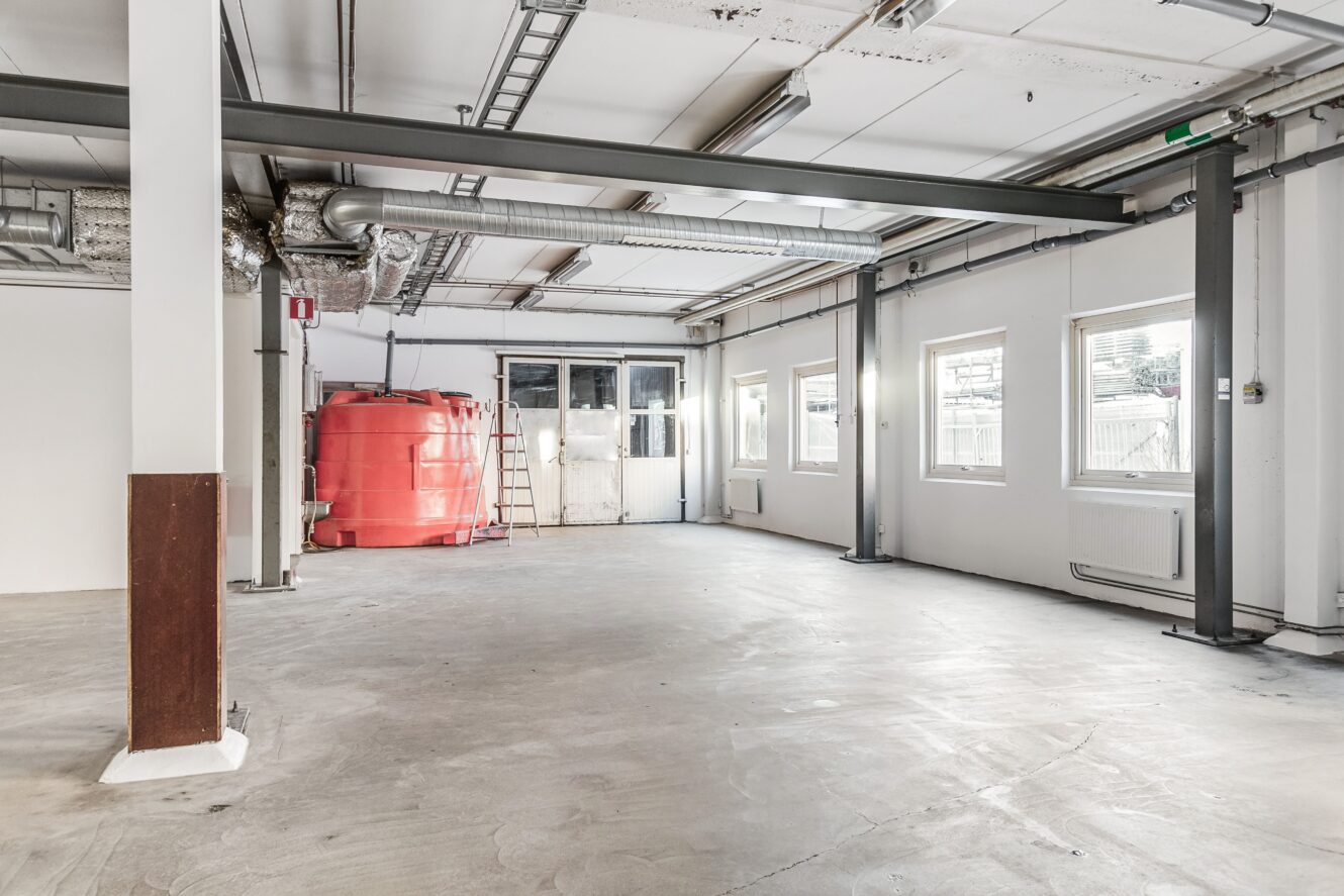En tom industrilokal med betonggolv, synliga kanaler, en röd tank i hörnet, en stege och flera fönster längs ena väggen.