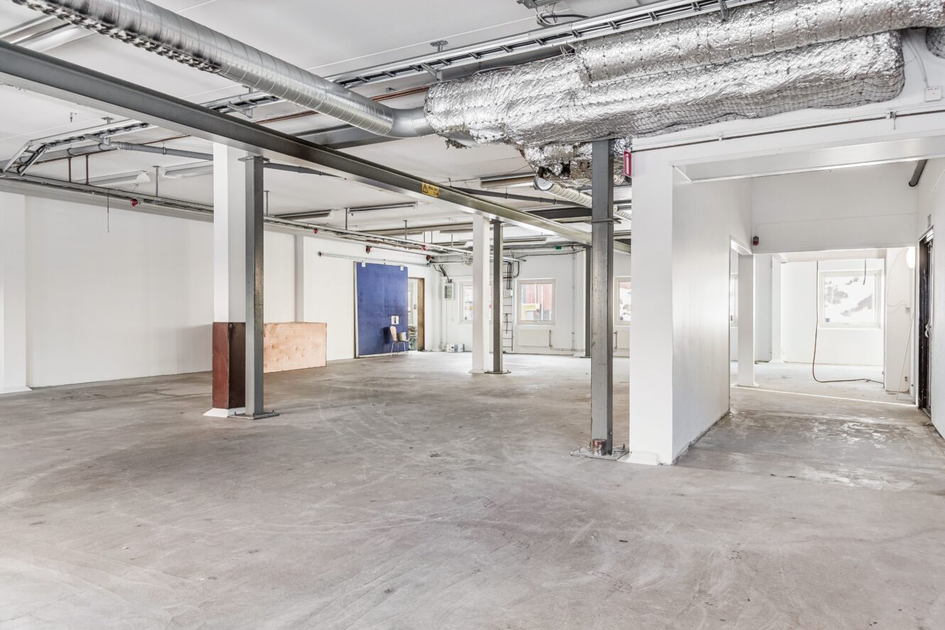 Rymligt, tomt rum i industriell stil med synliga kanaler och metallbalkar, ofärdiga golv, vita väggar och några små fönster.
