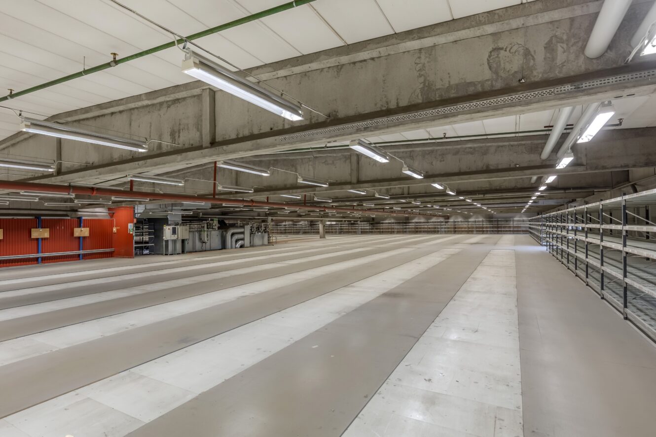 Stort, tomt industrilager med betongbalkar, stark takbelysning och markerade golvsektioner. Röd vägg med rör och utrustning synlig på vänster sida.