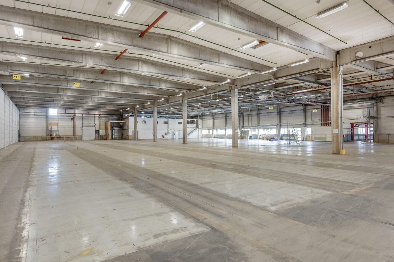 Rymlig, tom industrilagerlokal med högt i tak, betonggolv och synliga balkar, upplyst av lysrör.