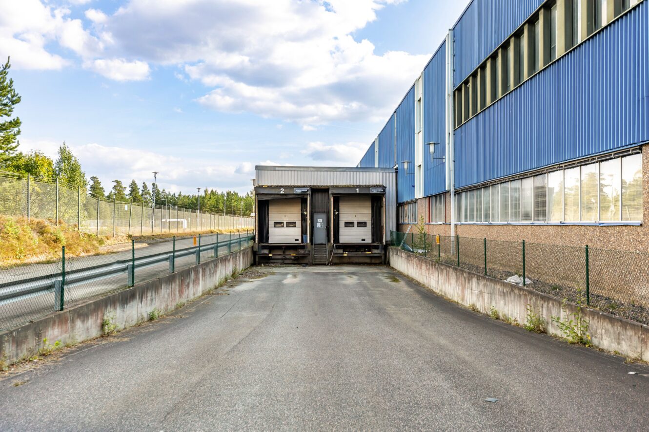 Två lastkajer på baksidan av ett blått industriellt lager, med en tom asfalterad uppfart och ett kedjelänkstaket till vänster.