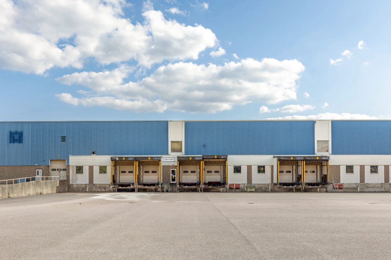 En stor blå lagerlokal med fem lastkajer och stängda dörrar, belägen under en delvis molnig himmel med en tom asfaltsyta framför.