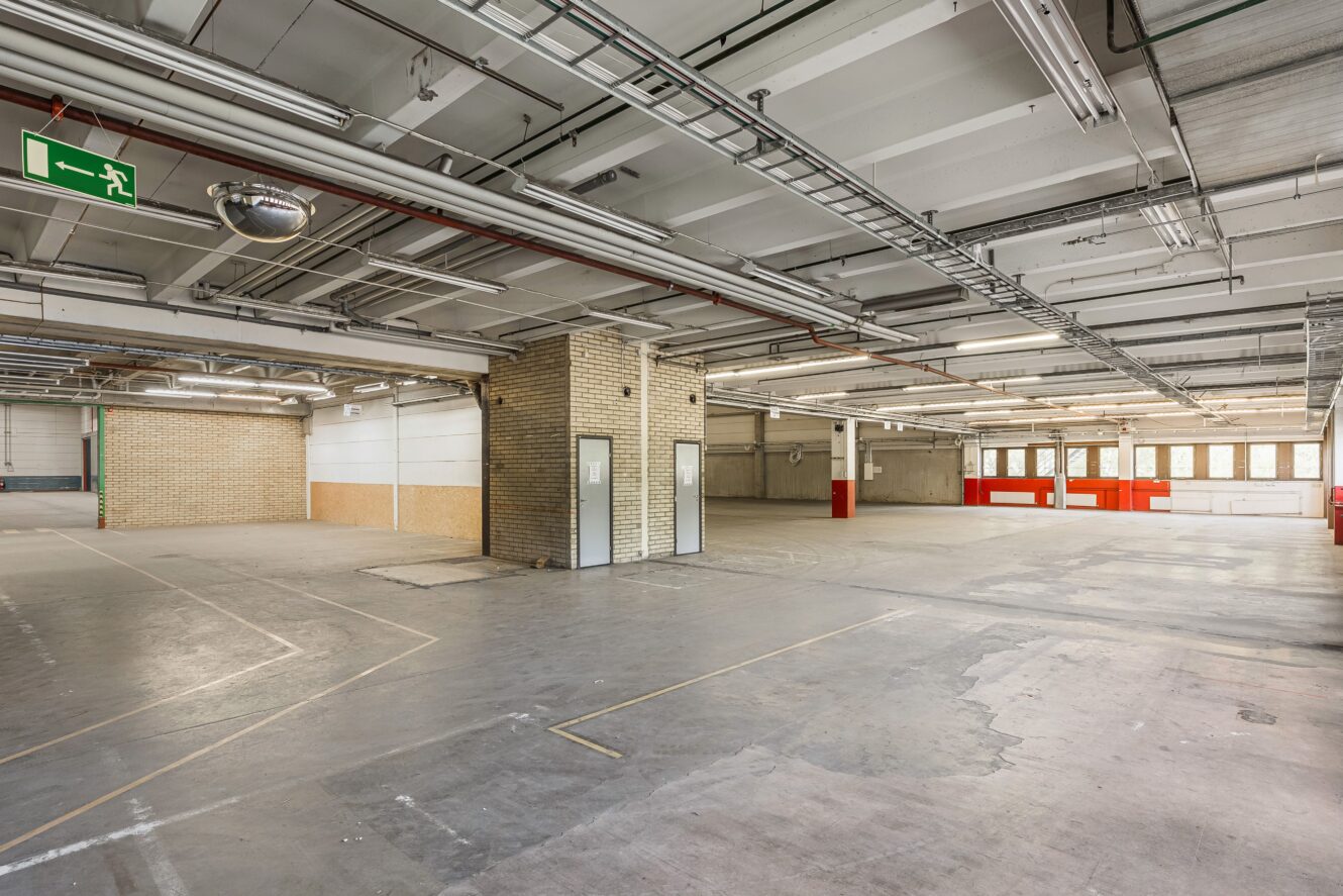 Rymlig, tom industrilokal med betonggolv, synliga tak, tegelpelare och stora fönster som släpper in naturligt ljus.