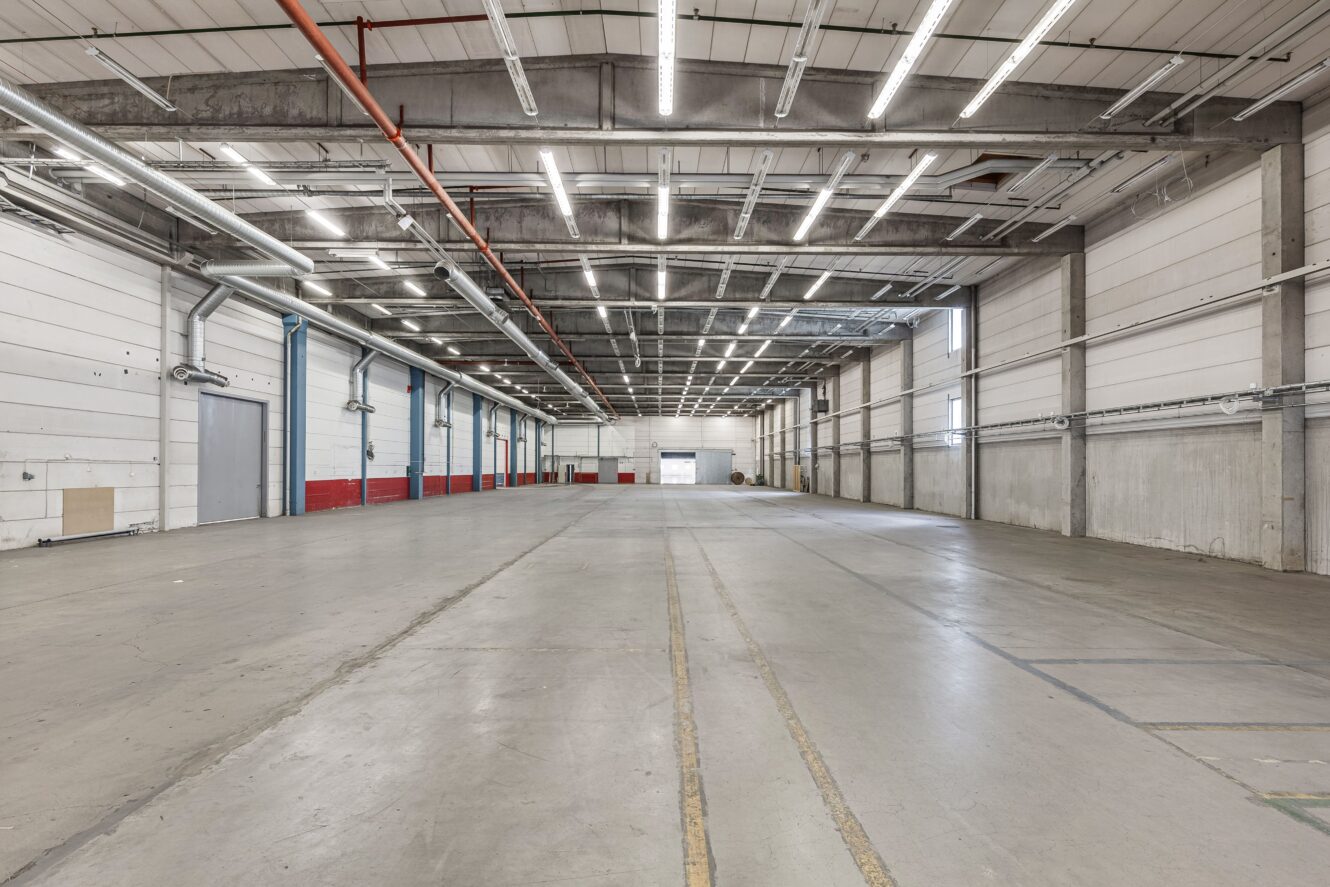 Rymlig, tom industrilagerinredning med högt i tak, synliga rör, takbelysning och betonggolv.