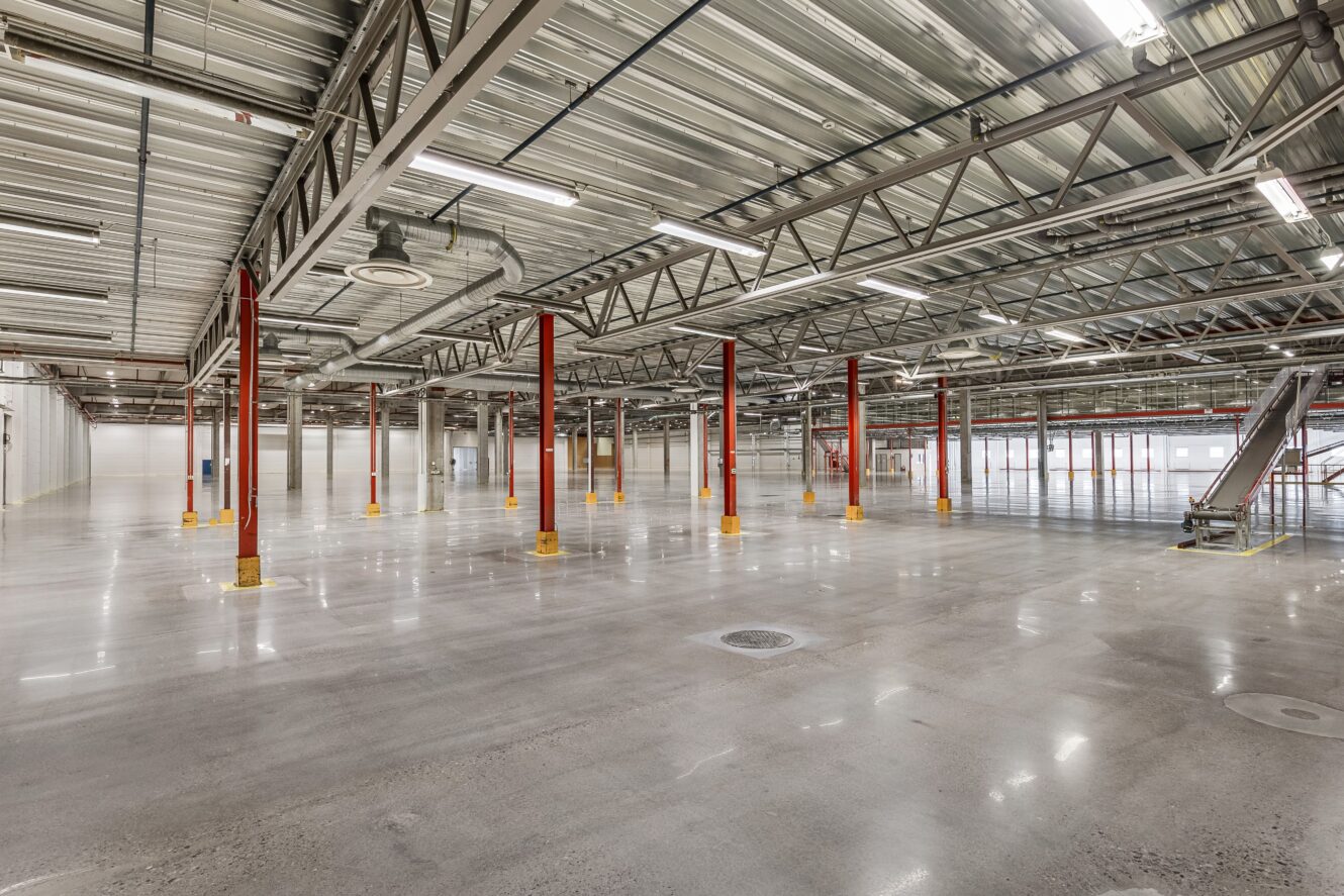Stort tomt industrilager med polerade betonggolv, röda stödpelare, synliga takbjälkar och gott om takbelysning.