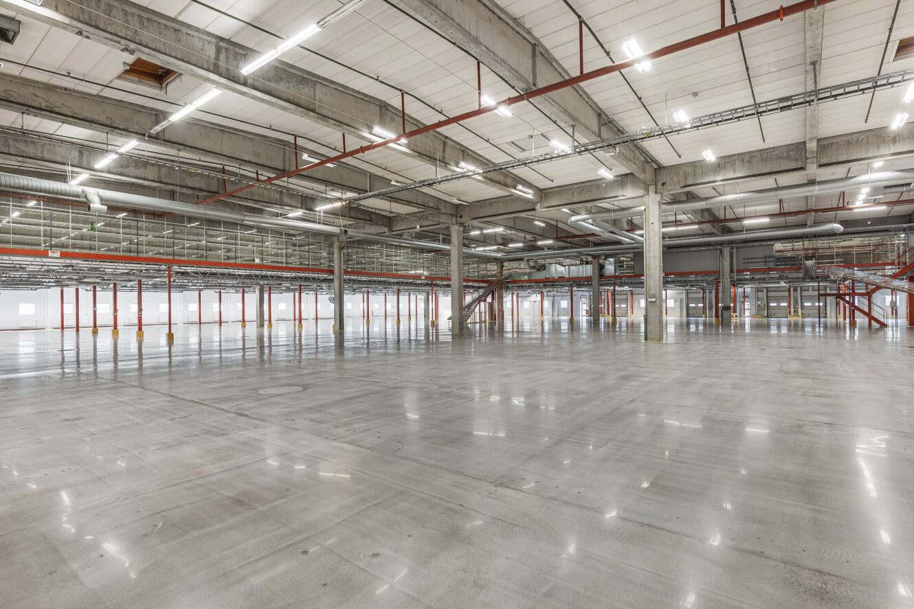 Stort, tomt industrilager med polerade betonggolv, synliga takkonstruktioner, takbelysning och metalltrappor längs sidoväggarna.