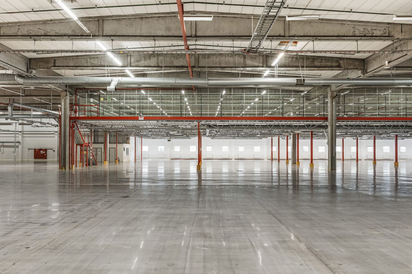 Stor, tom industrilagerinredning med betonggolv, synliga takbjälkar, röda stödpelare och rader av fönster som släpper in naturligt ljus.